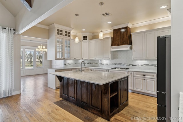 kitchen with light wood finished floors, a center island with sink, fridge, premium range hood, and backsplash