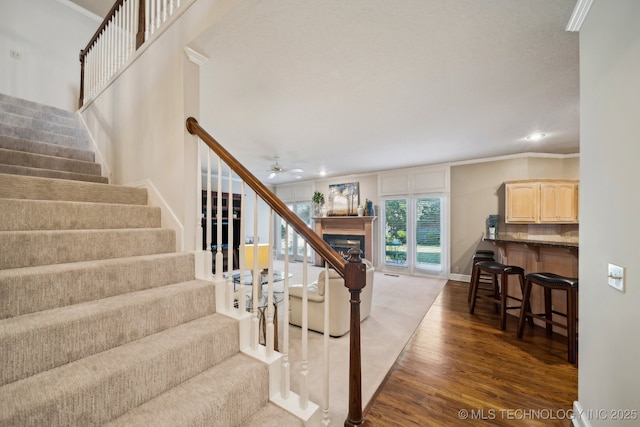 stairs featuring ceiling fan, a fireplace, wood finished floors, baseboards, and crown molding