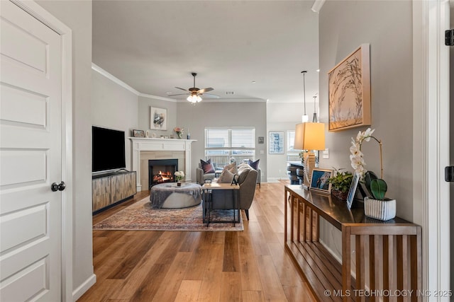 living room with a warm lit fireplace, baseboards, a ceiling fan, light wood-style flooring, and crown molding