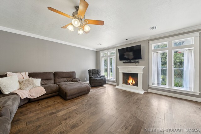 living area with a warm lit fireplace, visible vents, a ceiling fan, wood finished floors, and crown molding