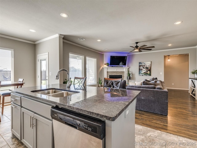 kitchen with a fireplace, stainless steel dishwasher, a kitchen island with sink, a sink, and dark stone counters