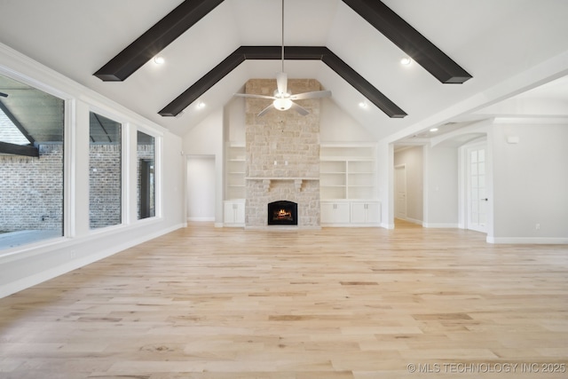 unfurnished living room with ceiling fan, beamed ceiling, a stone fireplace, light wood-style floors, and built in shelves