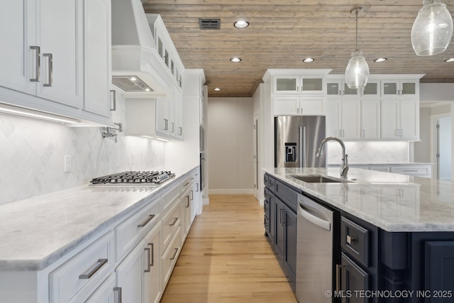 kitchen with a center island with sink, custom exhaust hood, stainless steel appliances, white cabinetry, and a sink