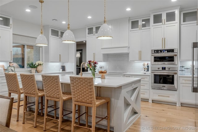 kitchen featuring double oven, a center island with sink, light countertops, and backsplash