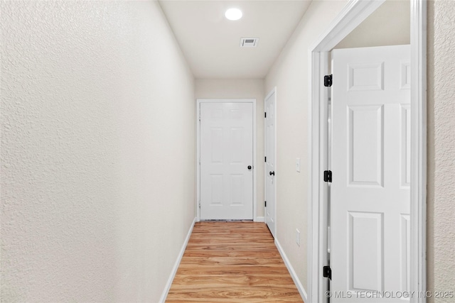 hallway with a textured wall, light wood finished floors, visible vents, and baseboards