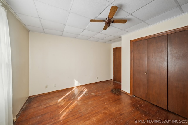 unfurnished bedroom featuring a closet, hardwood / wood-style floors, a ceiling fan, a drop ceiling, and baseboards