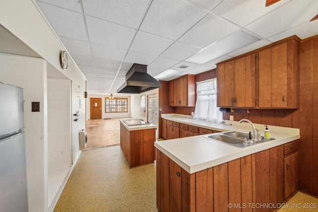 kitchen with wall chimney exhaust hood, brown cabinets, freestanding refrigerator, stovetop, and a sink