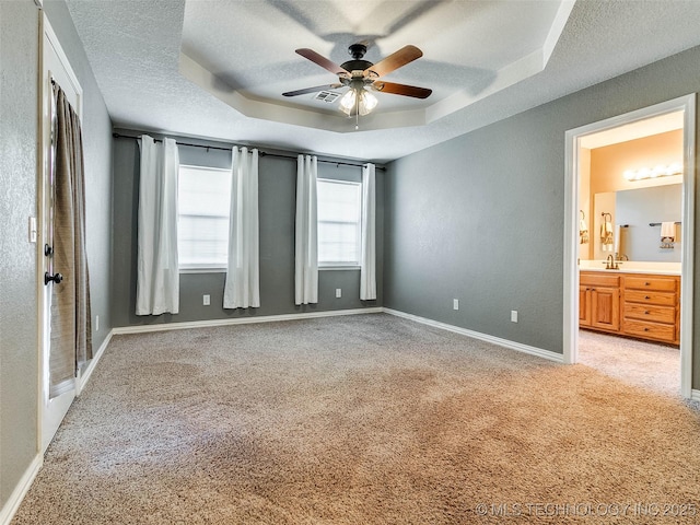 unfurnished bedroom featuring a textured ceiling, connected bathroom, light colored carpet, baseboards, and a raised ceiling