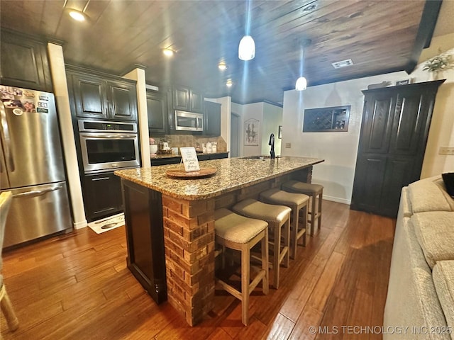 kitchen featuring a breakfast bar area, stainless steel appliances, dark wood-type flooring, an island with sink, and light stone countertops