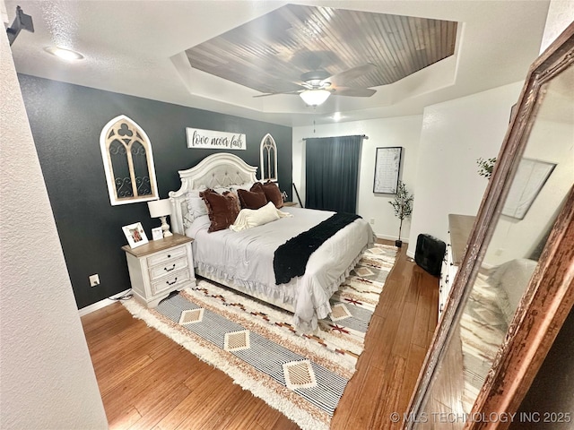 bedroom featuring a raised ceiling, a textured wall, a ceiling fan, wood finished floors, and baseboards