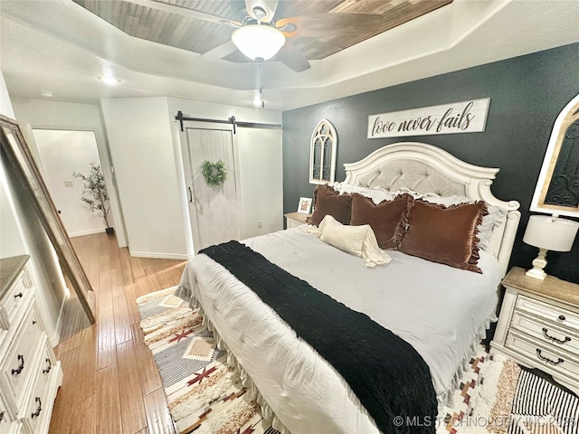 bedroom featuring light wood finished floors, a barn door, baseboards, a raised ceiling, and ceiling fan