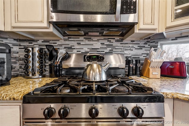 kitchen featuring light stone counters, cream cabinets, stainless steel appliances, decorative backsplash, and glass insert cabinets