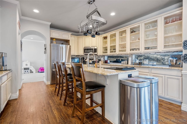 kitchen with a center island with sink, arched walkways, decorative backsplash, appliances with stainless steel finishes, and cream cabinetry