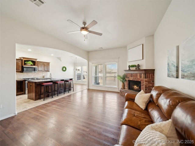 living room with arched walkways, light wood-style flooring, visible vents, a ceiling fan, and a brick fireplace