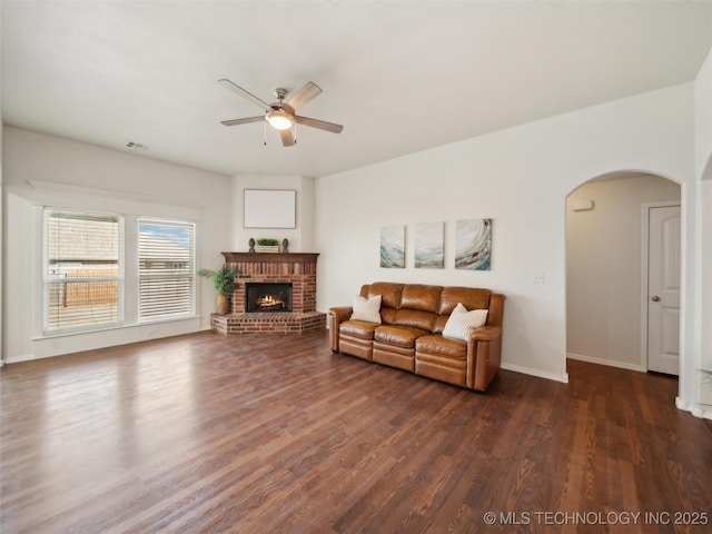 living room with dark wood-type flooring, arched walkways, a brick fireplace, and a ceiling fan