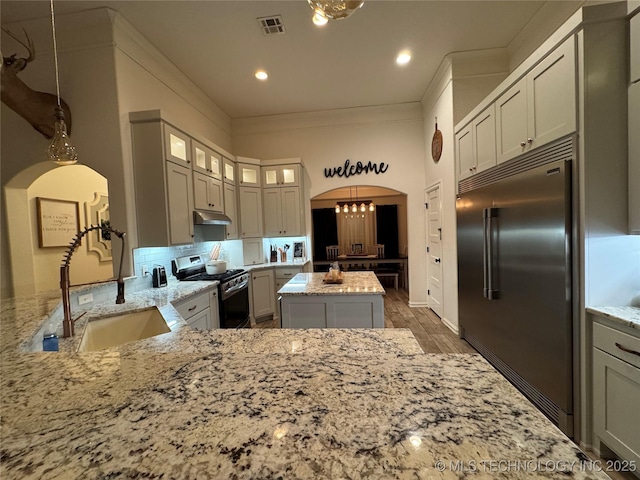 kitchen with arched walkways, light stone counters, under cabinet range hood, a sink, and appliances with stainless steel finishes