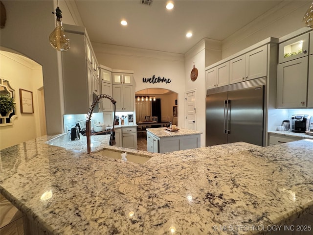 kitchen with arched walkways, built in fridge, a peninsula, ornamental molding, and light stone countertops