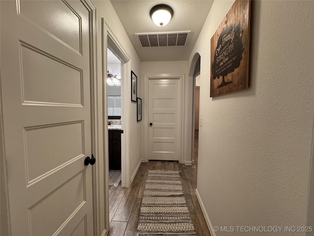 corridor with arched walkways, a textured wall, visible vents, baseboards, and dark wood-style floors