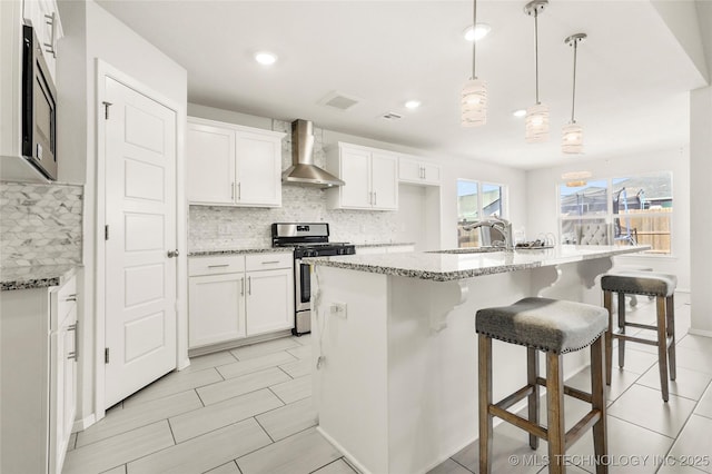 kitchen featuring light stone counters, appliances with stainless steel finishes, white cabinets, a sink, and wall chimney range hood