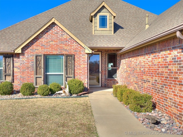 view of exterior entry with brick siding, a shingled roof, and a yard