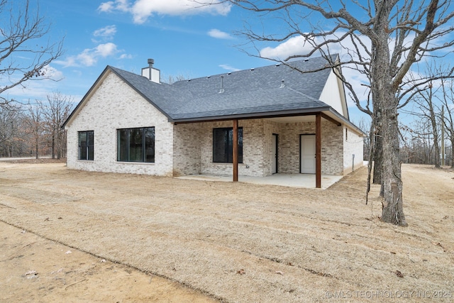 back of house with a shingled roof, a patio area, brick siding, and a chimney