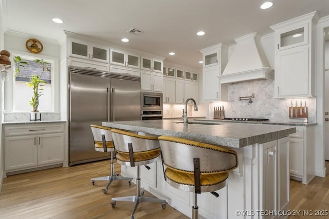 kitchen featuring visible vents, custom range hood, built in appliances, light wood-type flooring, and a sink