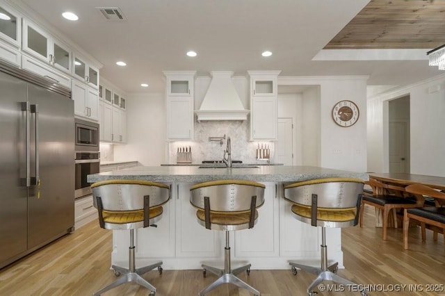 kitchen with built in appliances, light wood-style flooring, premium range hood, visible vents, and backsplash