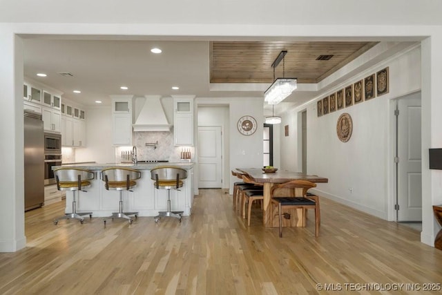 kitchen with a tray ceiling, custom range hood, light wood-style flooring, white cabinetry, and built in appliances