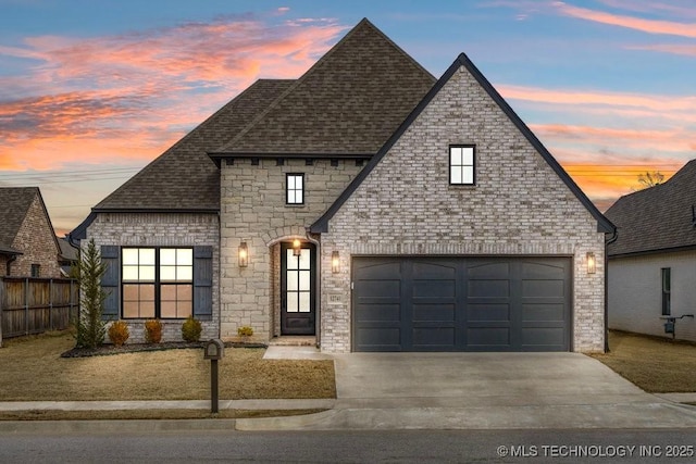 french country style house with driveway, brick siding, fence, and roof with shingles