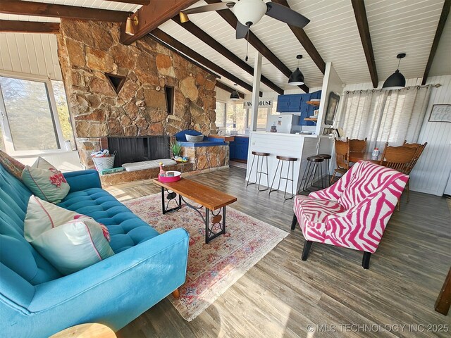 living area featuring vaulted ceiling with beams, a wealth of natural light, wood finished floors, and a stone fireplace