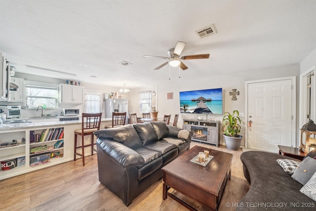 living room featuring light wood-style floors, visible vents, a ceiling fan, and a glass covered fireplace