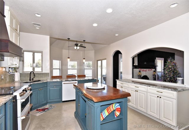 kitchen featuring blue cabinetry, butcher block counters, custom range hood, white appliances, and a peninsula