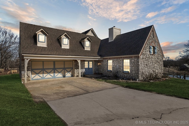 cape cod house with a garage, a shingled roof, driveway, a yard, and stone siding