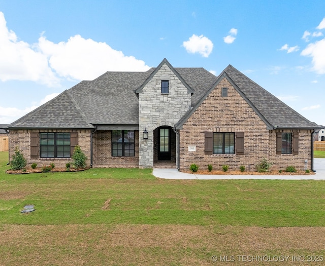 french country home featuring roof with shingles, a front lawn, and brick siding