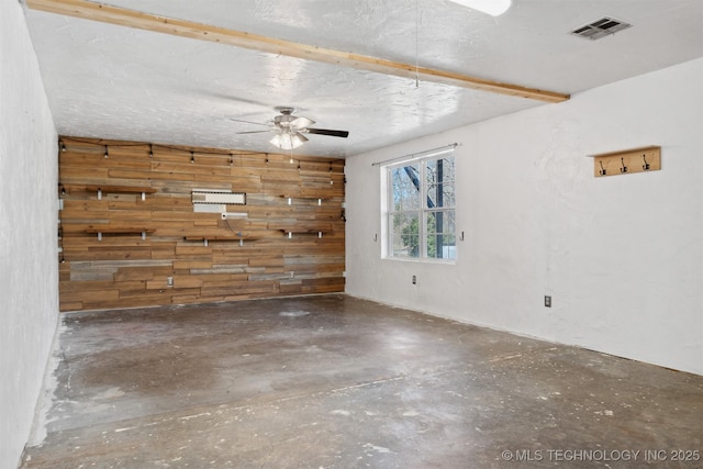 empty room featuring a textured ceiling, wood walls, a ceiling fan, visible vents, and unfinished concrete floors