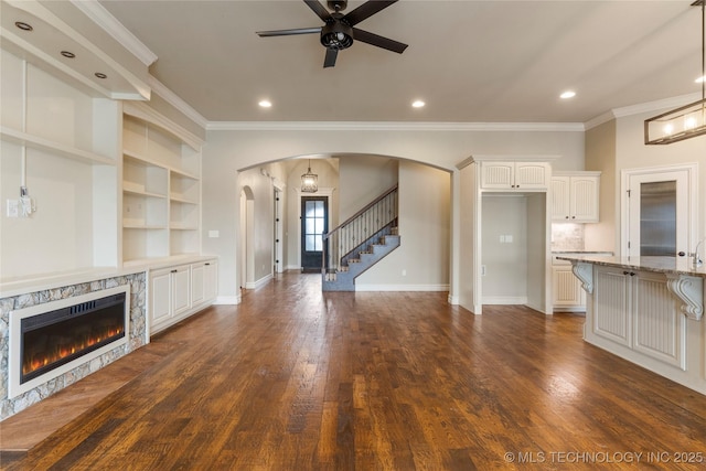 unfurnished living room featuring arched walkways, dark wood finished floors, baseboards, stairs, and a glass covered fireplace