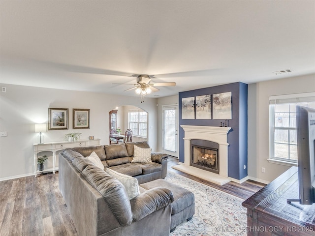 living area featuring a warm lit fireplace, baseboards, visible vents, arched walkways, and wood finished floors