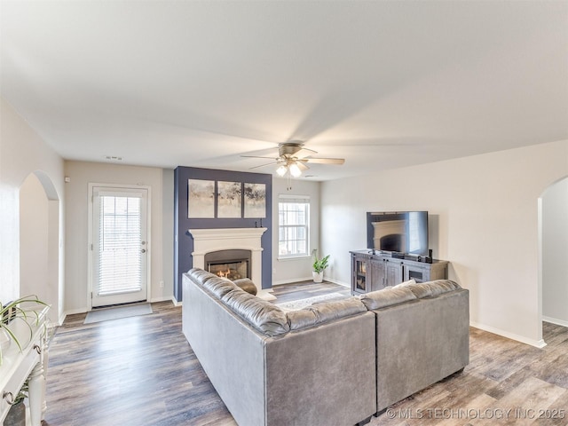 living area with baseboards, arched walkways, ceiling fan, wood finished floors, and a lit fireplace