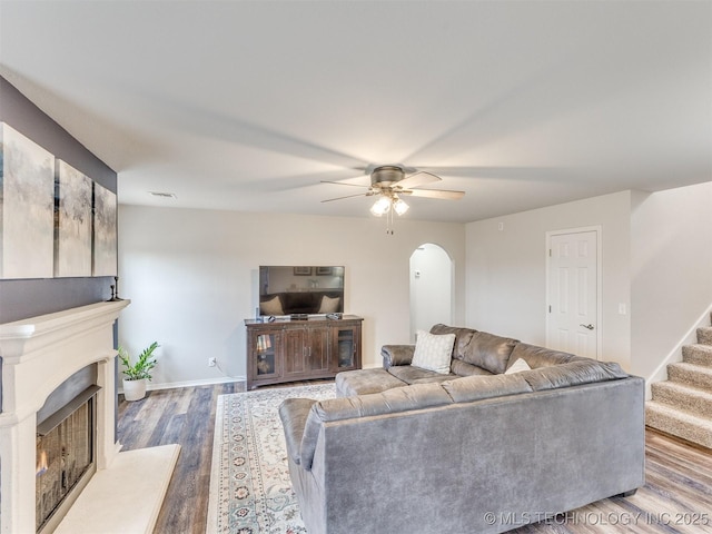 living room with arched walkways, a fireplace with raised hearth, a ceiling fan, wood finished floors, and stairs