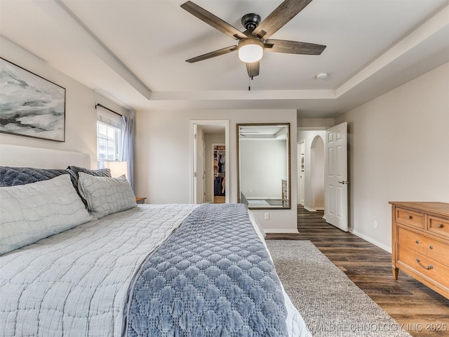 bedroom featuring arched walkways, wood finished floors, baseboards, a tray ceiling, and a walk in closet