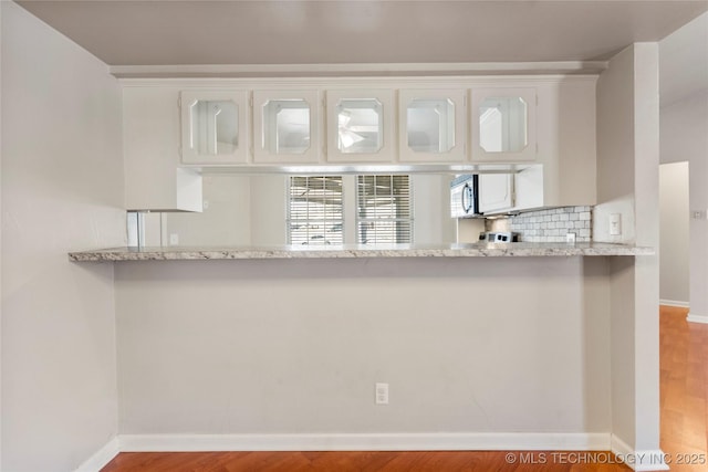 kitchen featuring a peninsula, white cabinets, decorative backsplash, stainless steel microwave, and glass insert cabinets