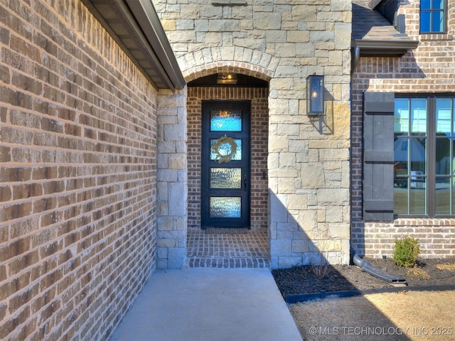 view of exterior entry with stone siding, brick siding, and roof with shingles