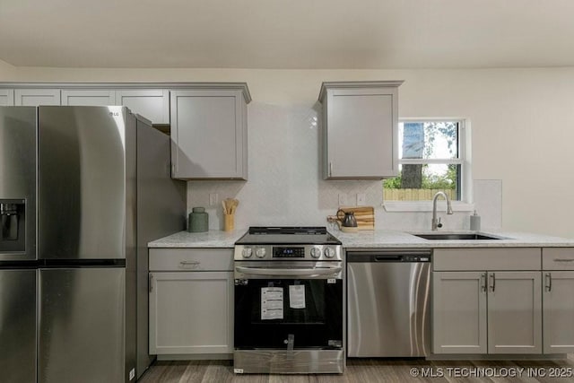 kitchen with light stone counters, gray cabinetry, a sink, appliances with stainless steel finishes, and decorative backsplash