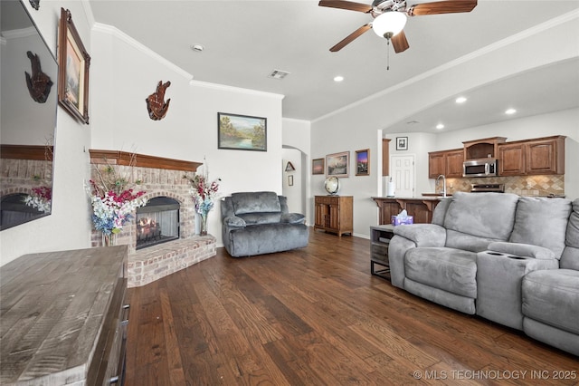 living room with dark wood-style floors, visible vents, arched walkways, crown molding, and a brick fireplace