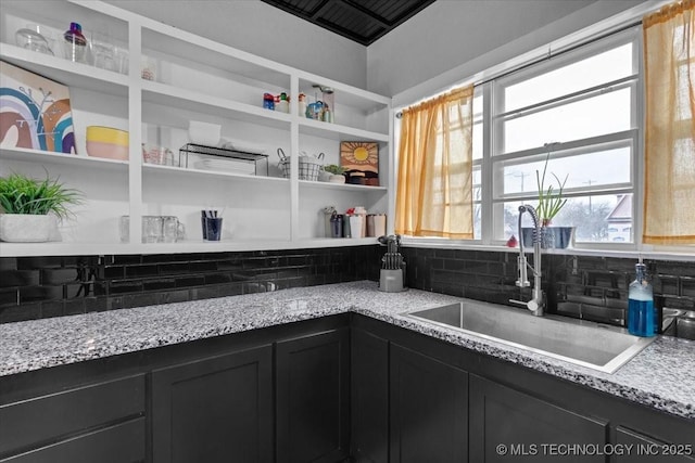 kitchen featuring decorative backsplash, light stone counters, dark cabinets, open shelves, and a sink