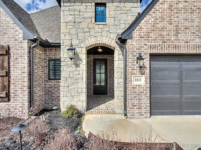 view of exterior entry featuring an attached garage, brick siding, stone siding, and roof with shingles