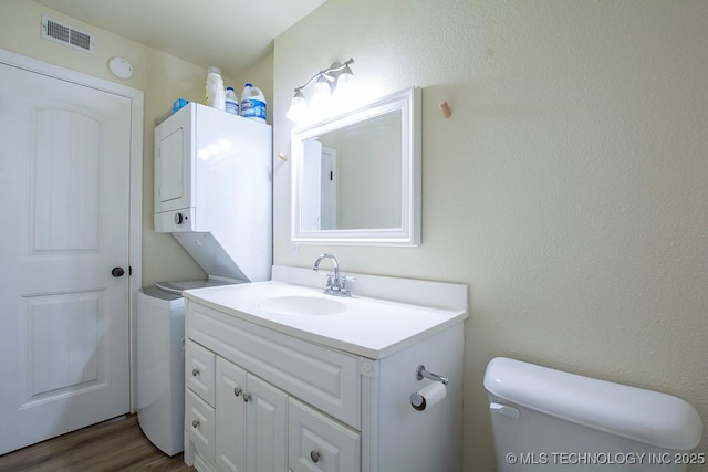 half bathroom with stacked washer and clothes dryer, visible vents, toilet, vanity, and wood finished floors