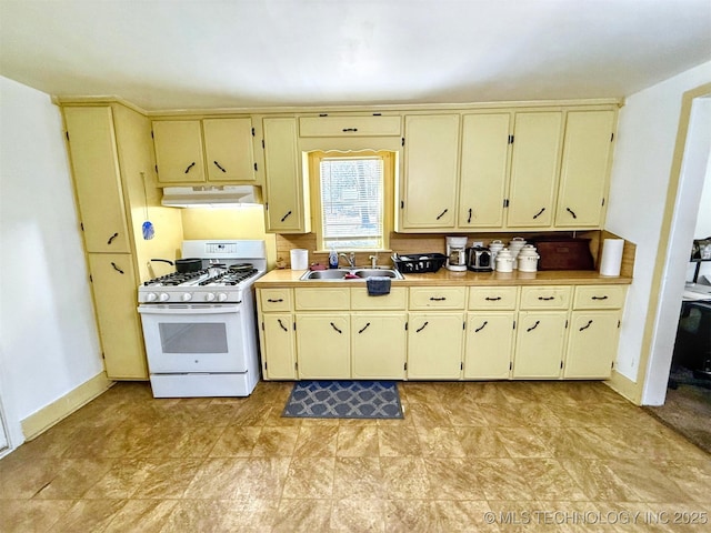 kitchen with cream cabinetry, a sink, under cabinet range hood, baseboards, and white gas range