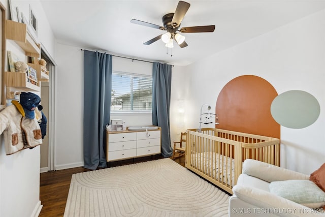 bedroom with ceiling fan, baseboards, a nursery area, and dark wood-style floors