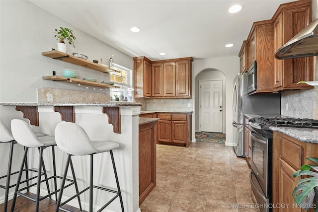 kitchen with wall chimney range hood, light stone countertops, a breakfast bar, brown cabinetry, and stainless steel appliances